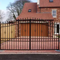 Kuta brama wjazdowa z czarnego metalu, ozdobiona ornamentami, przed domem z czerwonej cegły.