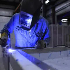 Spawanie aluminium migomatem. Pracownik w masce spawalniczej i niebieskiej koszuli precyzyjnie łączy metalowe elementy.
