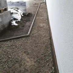 Żwirowa opaska wokół domu z kamienia, z ułożonymi paletami materiałów budowlanych po lewej stronie.