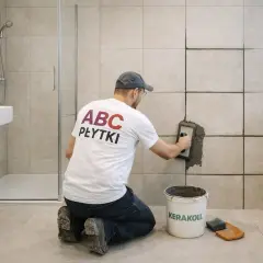 Mężczyzna w koszulce "ABC PŁYTKI" klęczy i fuguje ściany w łazience. Pokazuje, jak fugować płytki.
