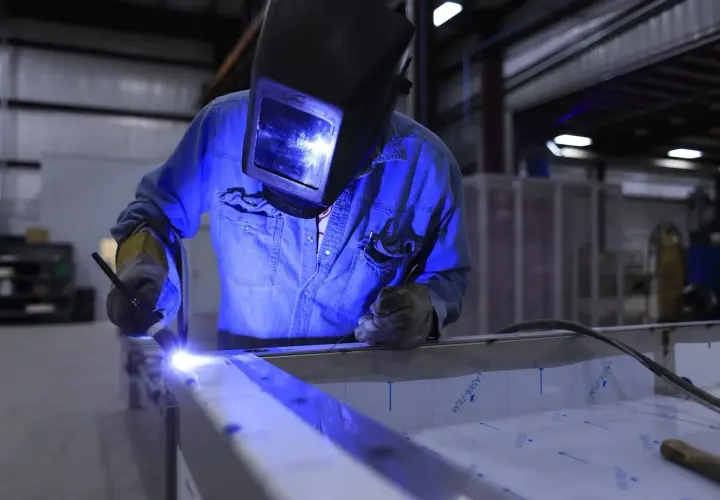 Spawanie aluminium migomatem. Pracownik w masce spawalniczej i niebieskiej koszuli precyzyjnie łączy metalowe elementy.