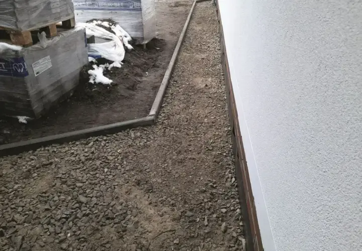 Żwirowa opaska wokół domu z kamienia, z ułożonymi paletami materiałów budowlanych po lewej stronie.
