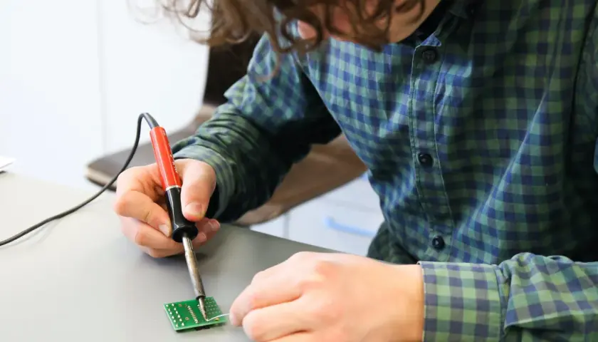 Młoda osoba w kratę wykonuje lutospawanie małej płytki drukowanej.