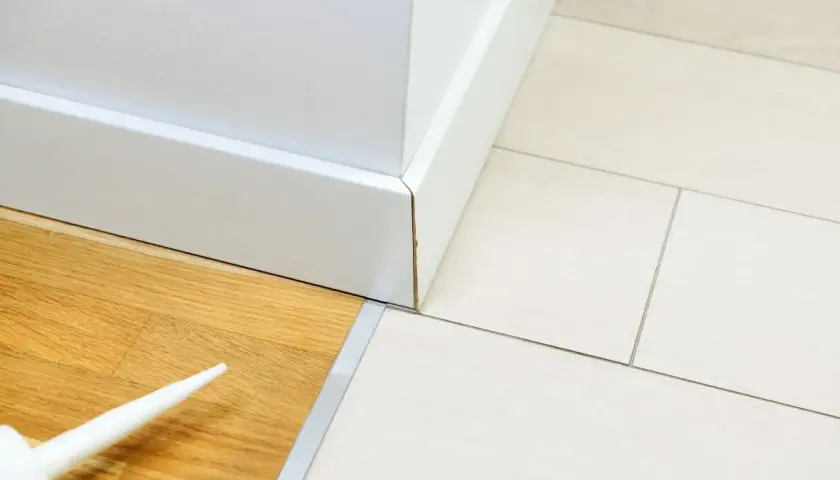 Biała listwa przypodłogowa łączy panele drewniane z jasnymi płytkami. Widać końcówkę tubki z silikonem, gotową do uszczelnienia szczelin.