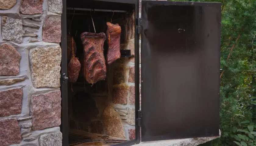 Prosta wędzarnia murowana z wędzonymi mięsami wiszącymi w środku. Drzwi otwarte, widać kamienną konstrukcję i zielone drzewa w tle.