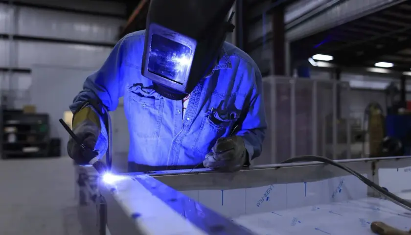 Spawanie aluminium migomatem. Pracownik w masce spawalniczej i niebieskiej koszuli precyzyjnie łączy metalowe elementy.