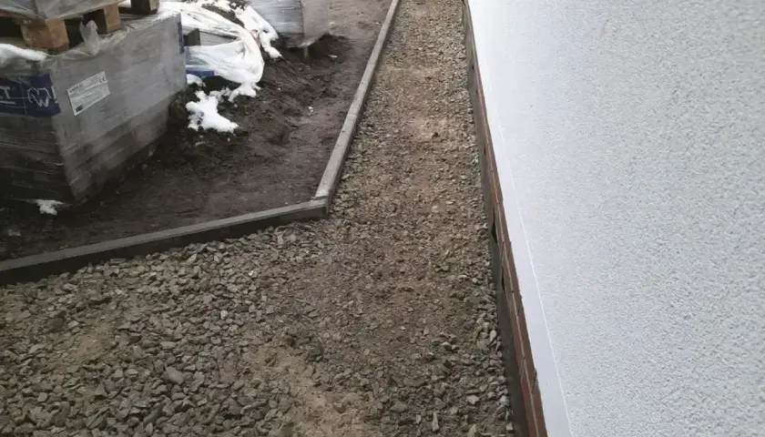 Żwirowa opaska wokół domu z kamienia, z ułożonymi paletami materiałów budowlanych po lewej stronie.