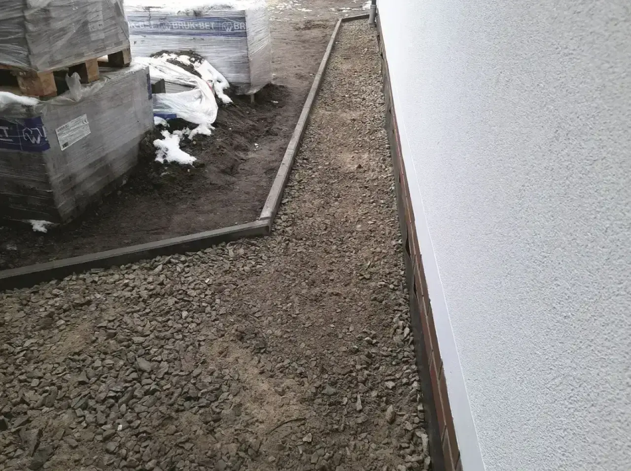 Żwirowa opaska wokół domu z kamienia, z ułożonymi paletami materiałów budowlanych po lewej stronie.
