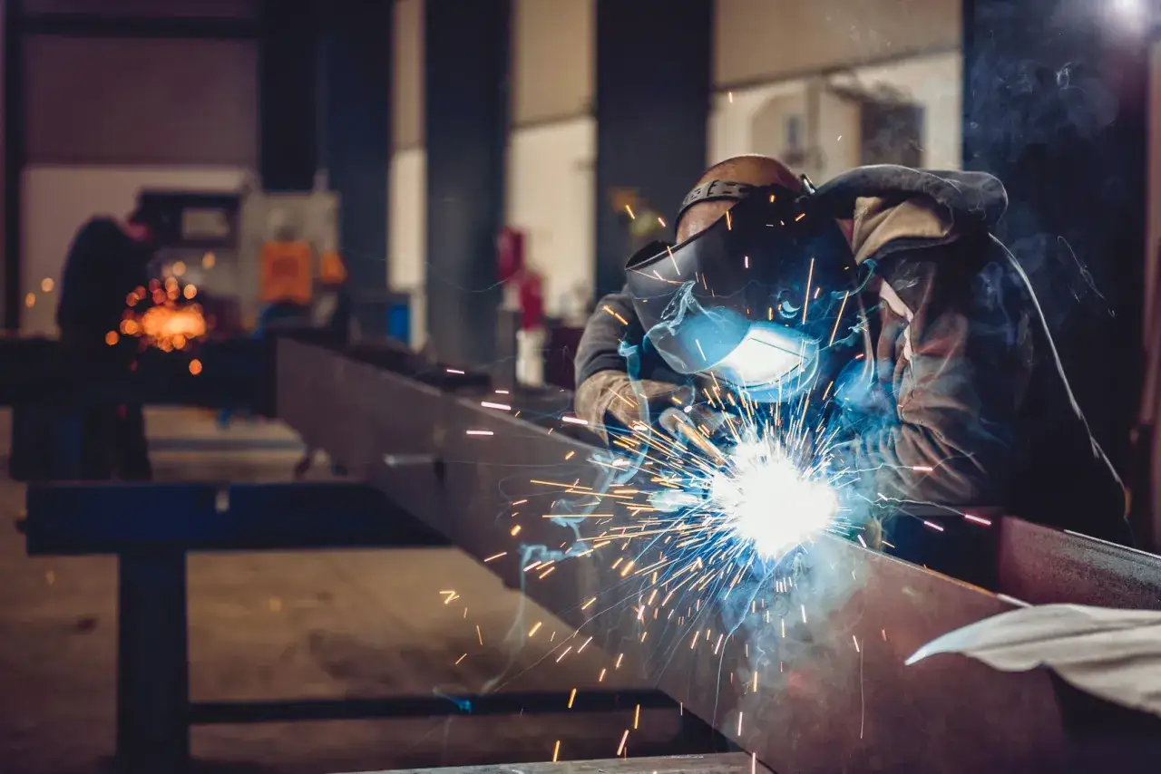 Spawacz TIG w masce ochronnej tworzy iskrzący łuk, łącząc metalowe elementy. W tle inny pracownik również spawa.